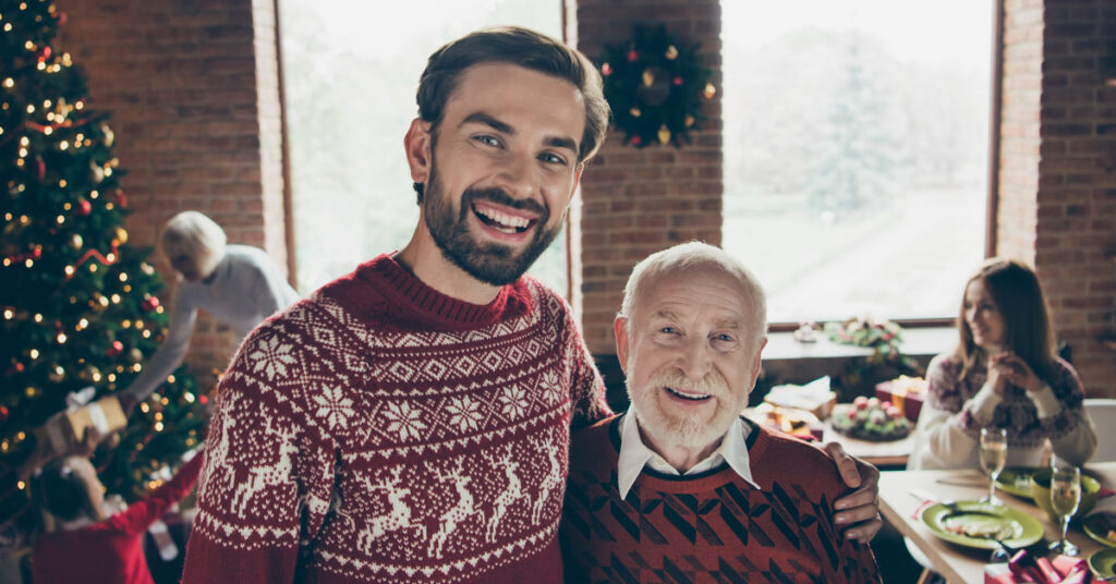 Elderly balding father and son with full head of hair wearing Christmas sweaters by the tree.