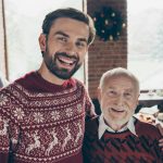 Elderly balding father and son with full head of hair wearing Christmas sweaters by the tree.